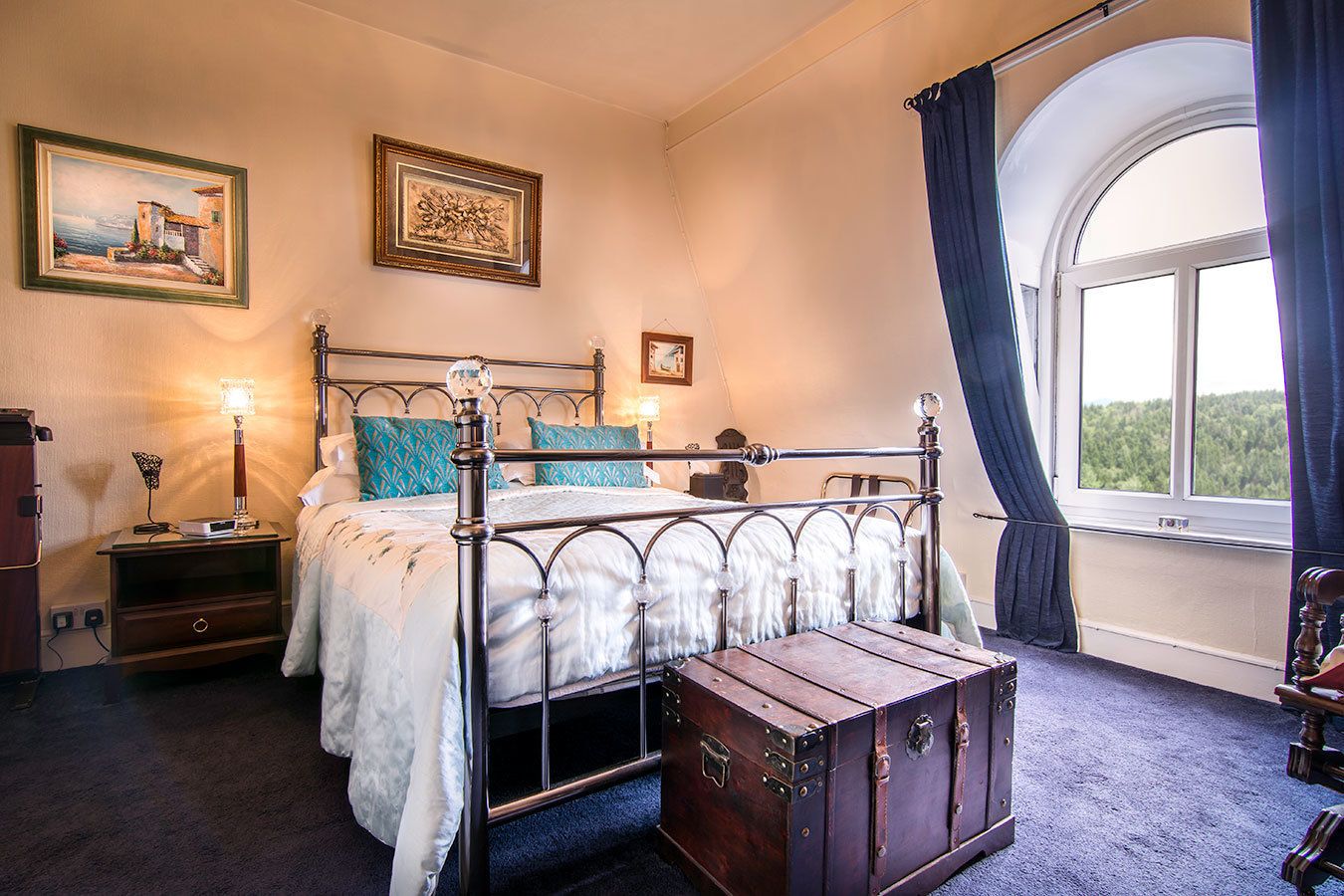 A bedroom with a metal bed frame, a wooden chest, two bedside tables, a lamp, and a window with curtains.