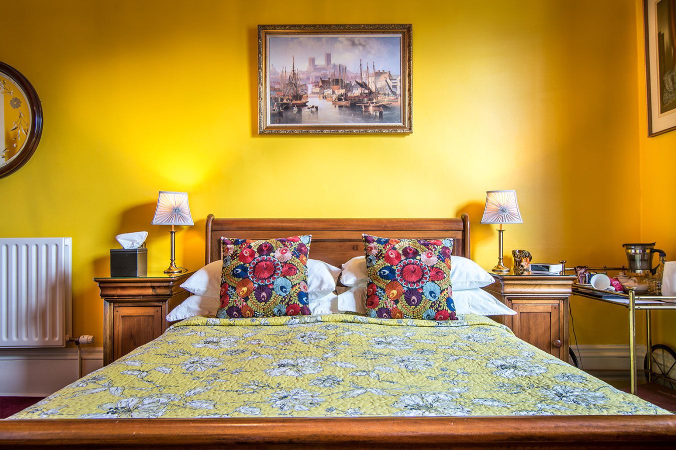 A bed with floral bedding, two pillows, bedside tables with lamps, a radiator, and a framed painting on the wall.