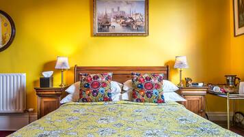 A bed with floral bedding, two pillows, bedside tables with lamps, a radiator, and a framed painting on the wall.