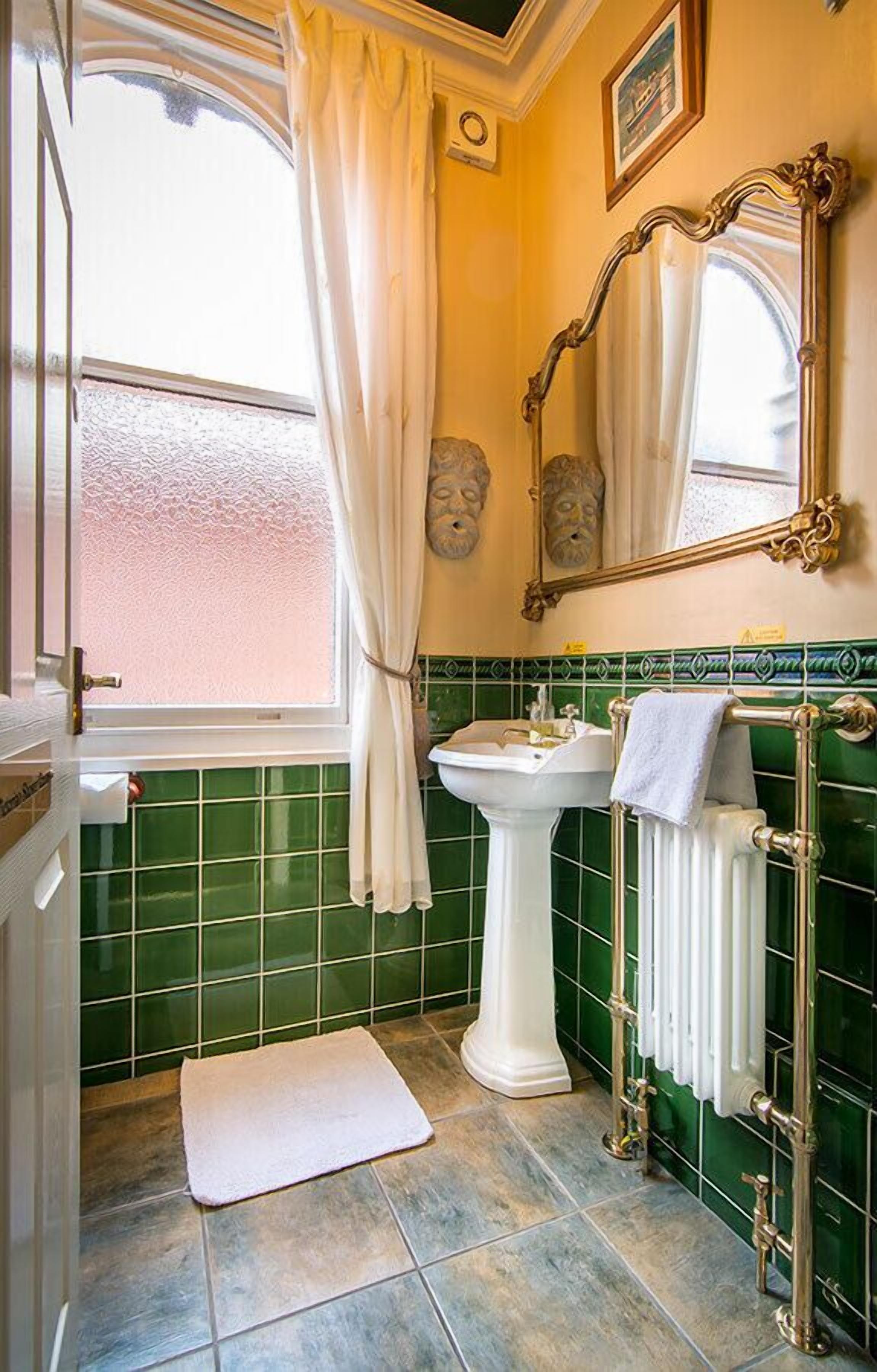 Un baño con lavamanos pedestal, un radiador, un perchero para toallas y una ventana con cortinas.