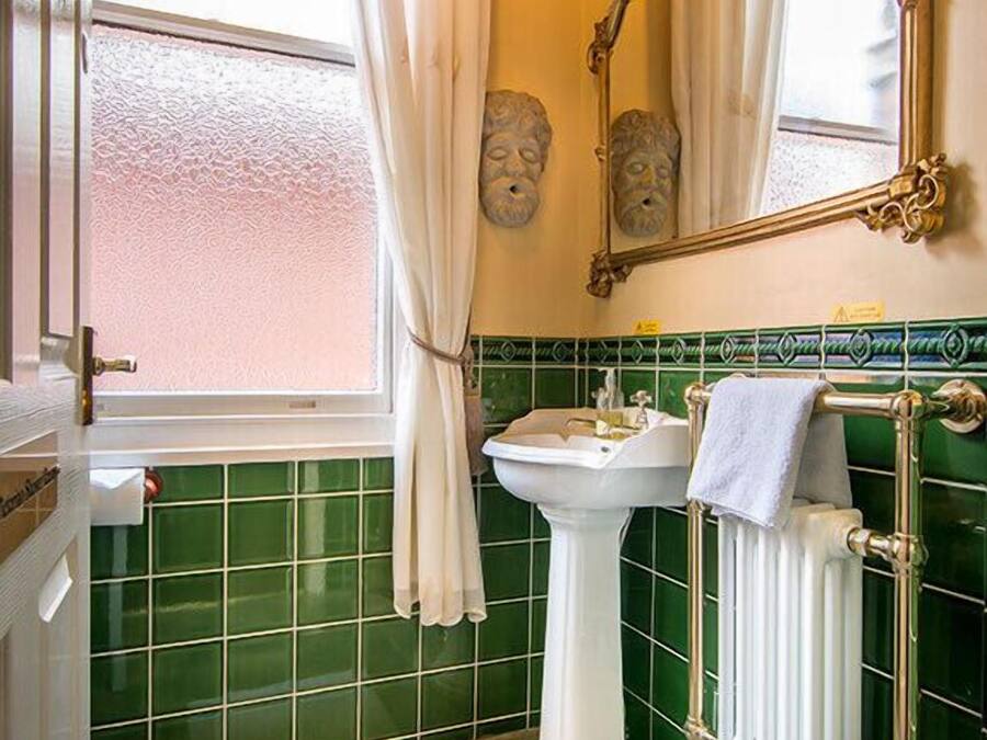 Un baño con lavamanos pedestal, un radiador, un perchero para toallas y una ventana con cortinas.