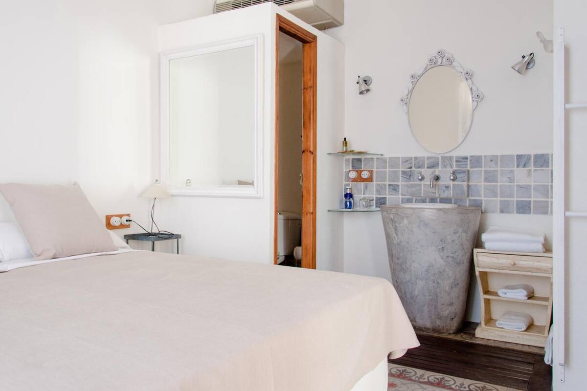 A bedroom with a bed, a bedside table, a lamp, a large mirror, a sink, and a towel rack.