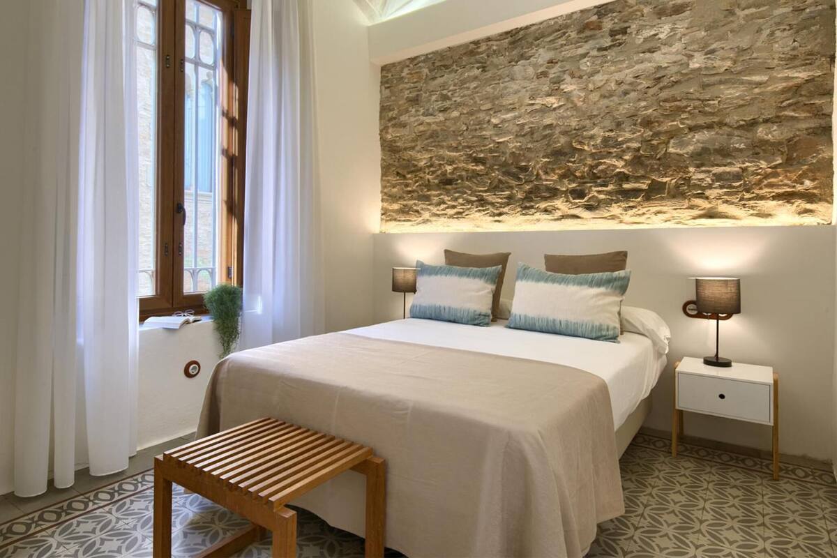 A bedroom with a bed, bedside tables, a bench, a stone wall, and a window with curtains.