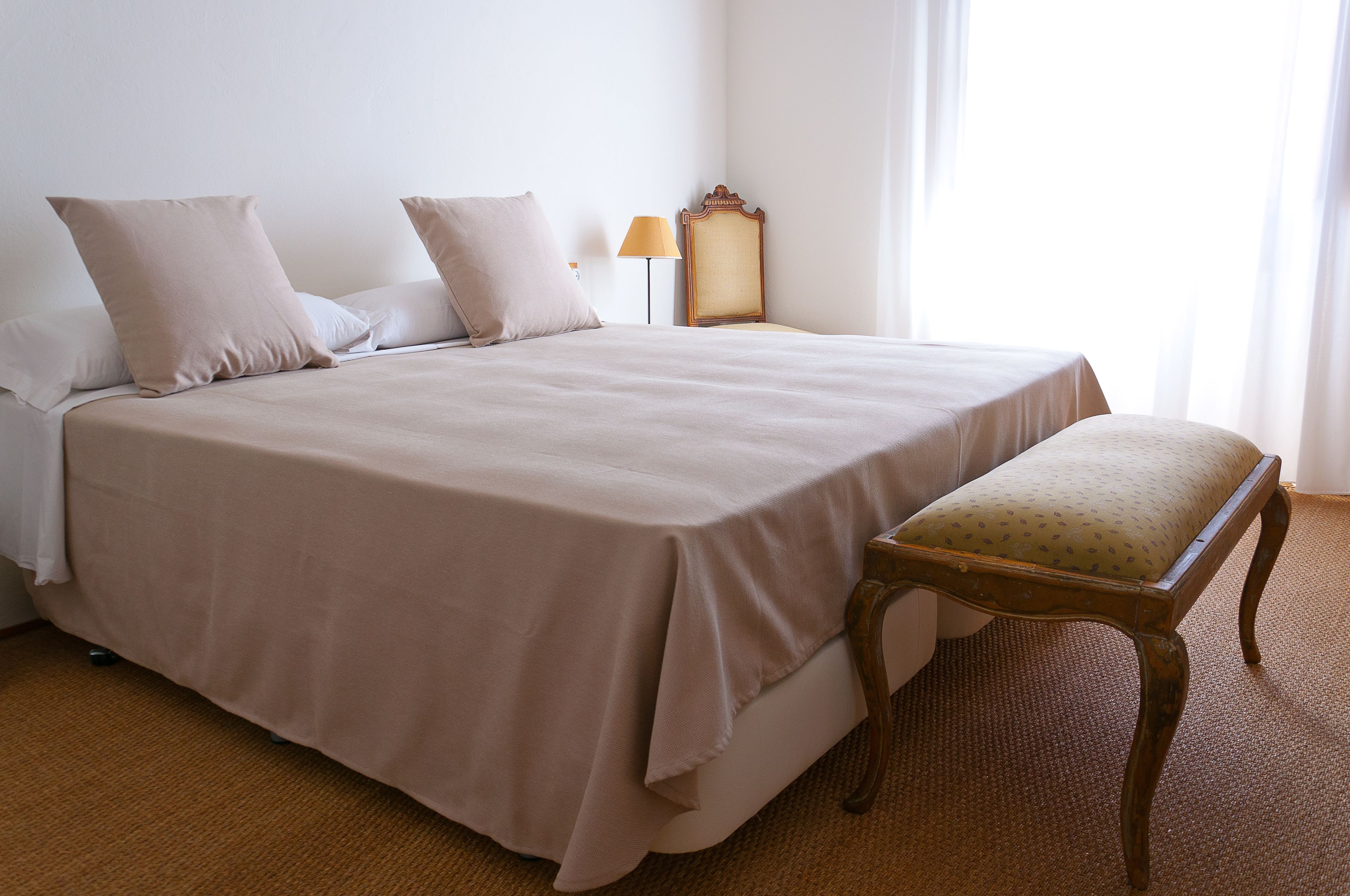 Una cama bien hecha con un banco a juego, una lámpara de noche y un espejo en la pared.