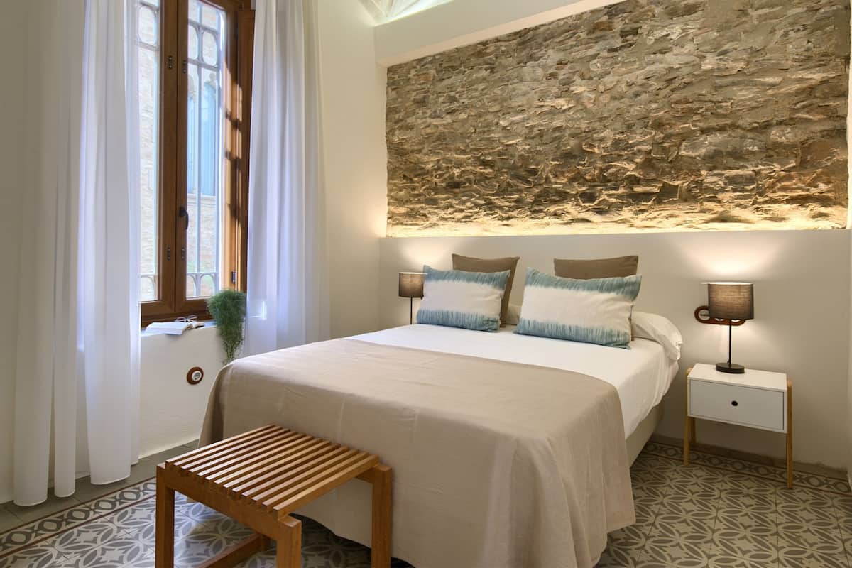 A bedroom with a bed, a wooden bench, a nightstand with a lamp, and a stone wall.