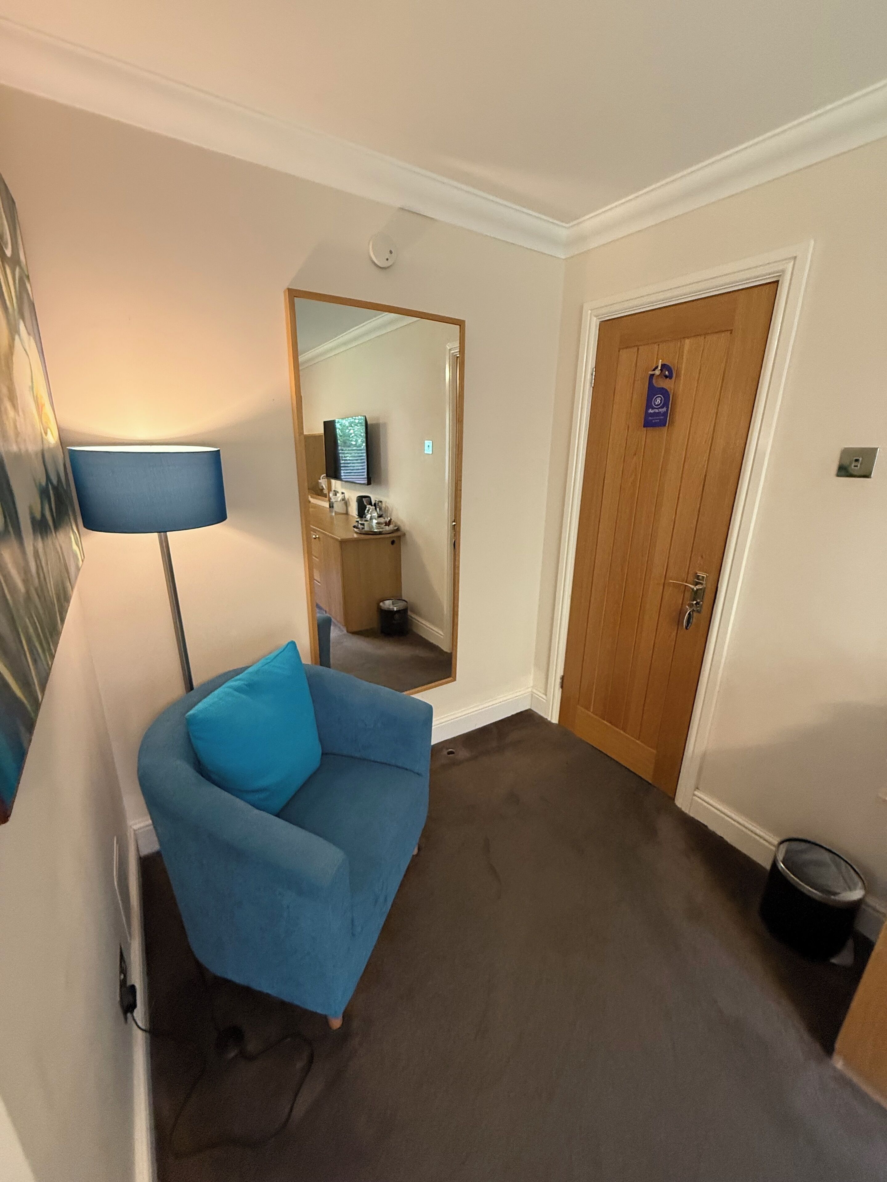 A hotel room with a blue sofa, a lamp, a wooden door, and a small trash bin.