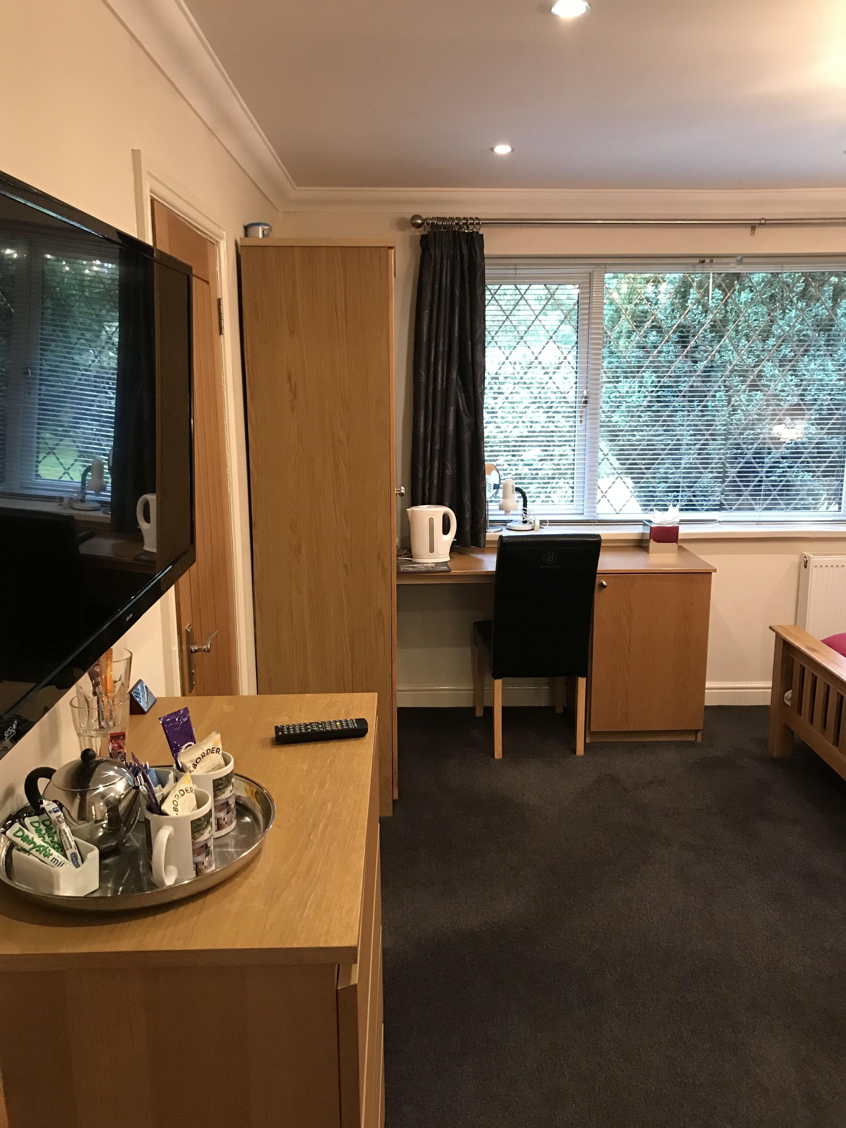 A compact room with a TV, a desk, a chair, a small cabinet, and a window with blinds.
