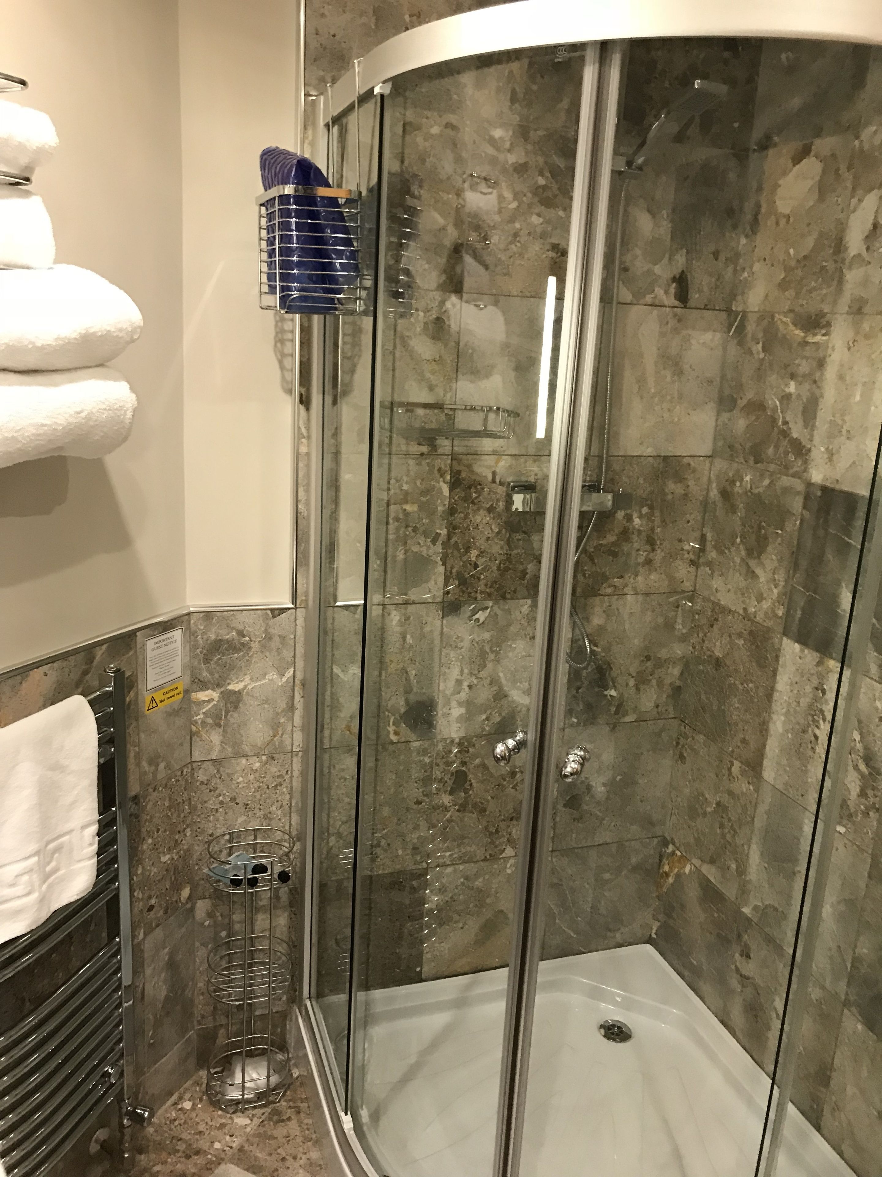 A glass-enclosed shower with a marble wall, a towel rack with white towels, and a metal shelf with toiletries.