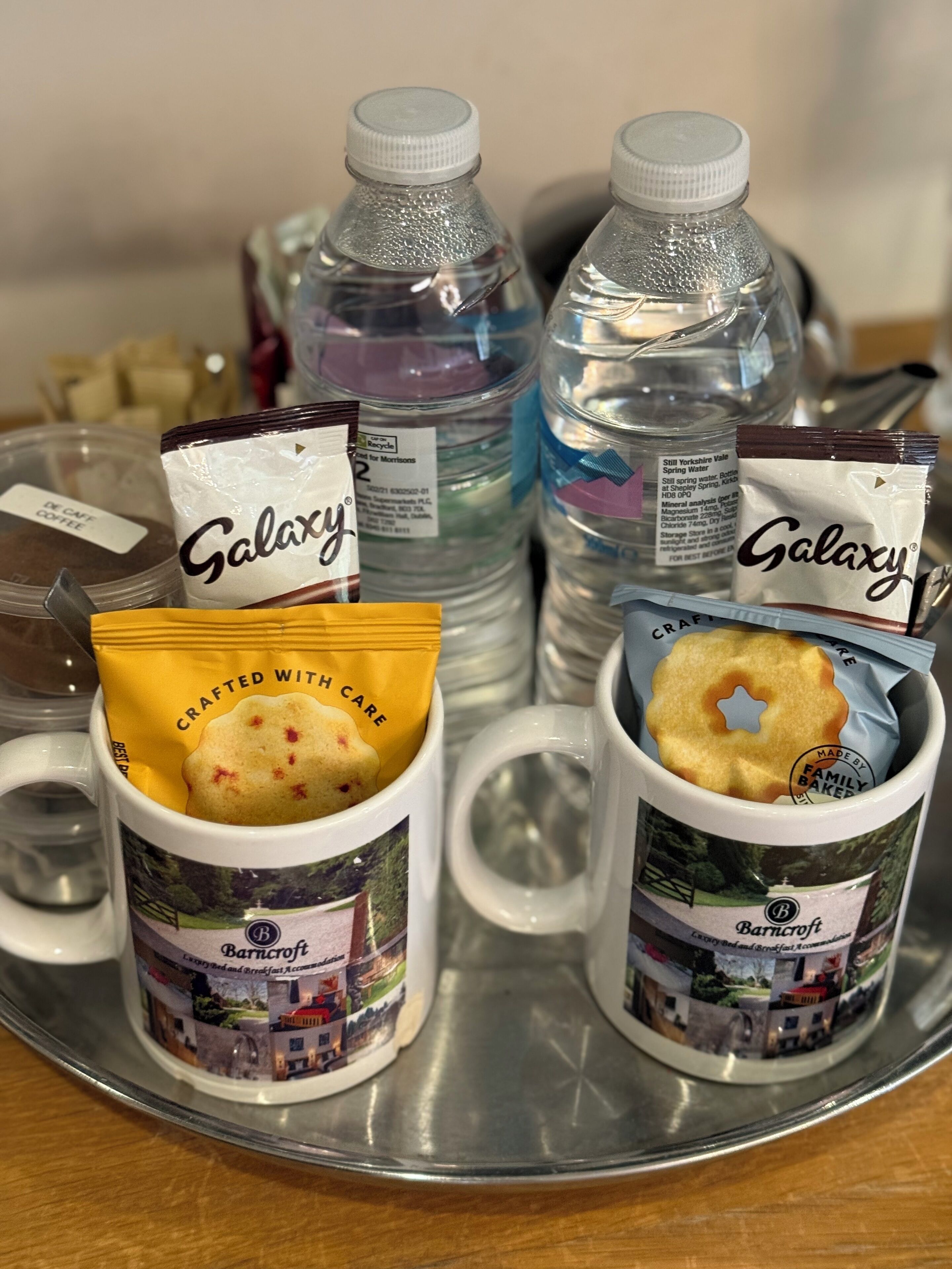 A tray with two mugs, each containing a pastry and a packet labeled "Galaxy". There are also two water bottles and a packet labeled "DX CAFE COFFEE".