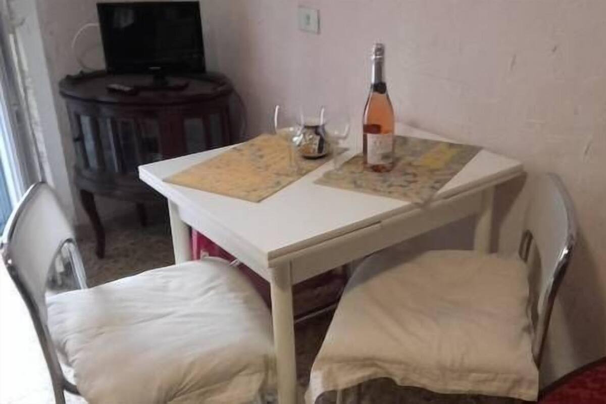 A small room with a white table, two white chairs, a bottle of wine, and a television in the background.