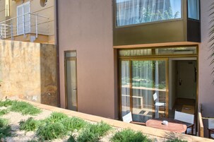 Un edificio moderno con puerta de entrada de vidrio, un pequeño patio con mesa y sillas, y vistas del entorno.