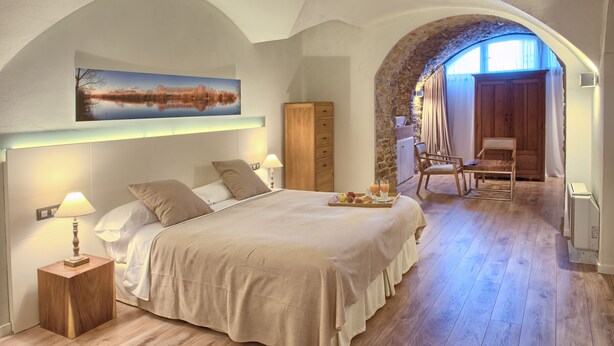 A bedroom with a large bed, a nightstand, a wooden wardrobe, and a dining table with a tray of food and drinks. - Hotel el Petit Convent (Begur)