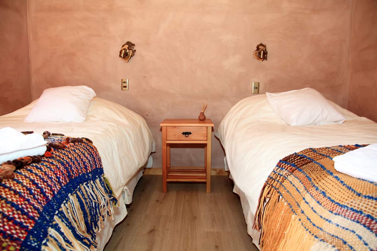 Ein Zimmer mit zwei Betten, jedes mit einem weißen Kopfkissen und einer farbenfrohen Decke. Dazwischen befindet sich ein kleiner Nachttisch aus Holz mit einer Schublade.