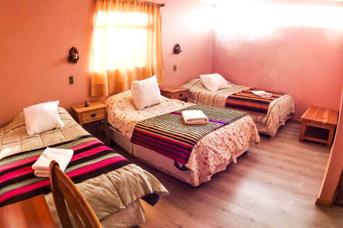 Ein Zimmer mit drei Betten, Holzboden und einem sichtbaren Badezimmer mit blauen Fliesen.