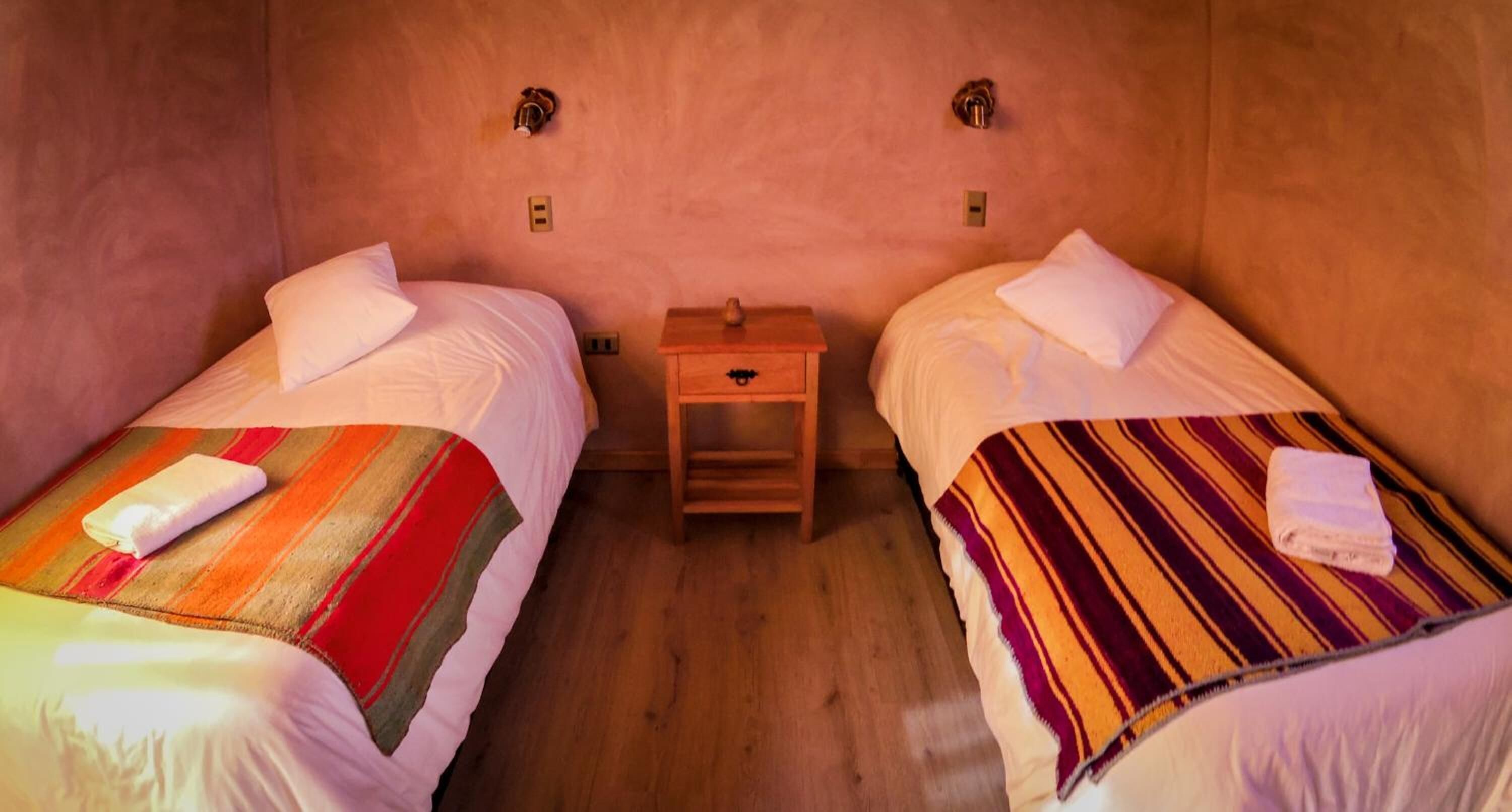 Une chambre avec deux lits, chacun recouvert d’une couverture à rayures et agrémenté d’un oreiller blanc. Une petite table de chevet en bois se trouve entre les deux lits, et un petit objet y est posé.