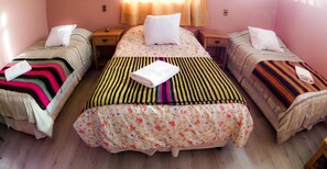 A bedroom with three single beds, each with a different patterned bedspread. There are two bedside tables with lamps, and a window with curtains.