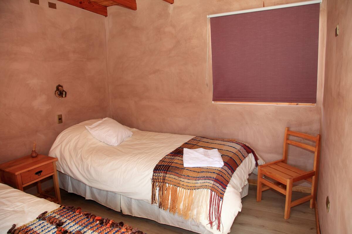 Ein Schlafzimmer mit einem Einzelbett, einem Nachttisch aus Holz, einem Stuhl und einem Fenster mit Jalousie.