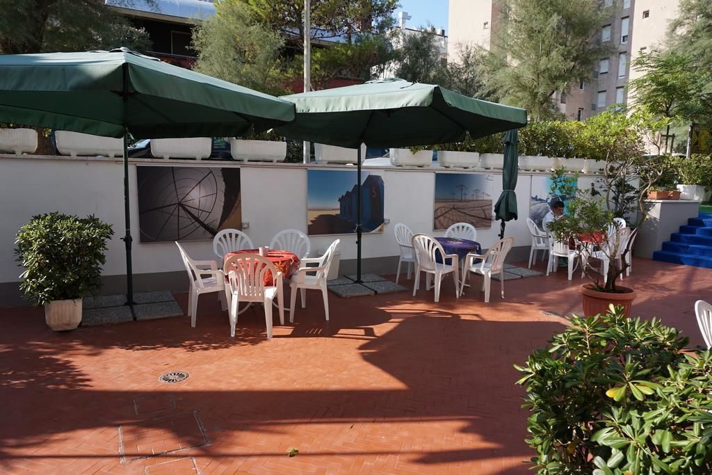 Un patio al aire libre con sillas y mesas de plástico blanco, sombrillas verdes y plantas en macetas.