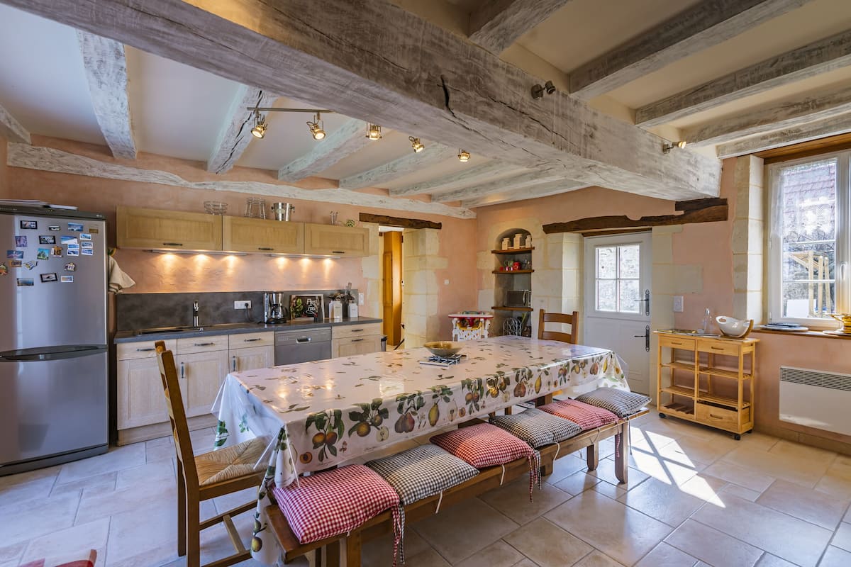A rustic kitchen with exposed beams, a dining table with benches, and a wooden cabinet.