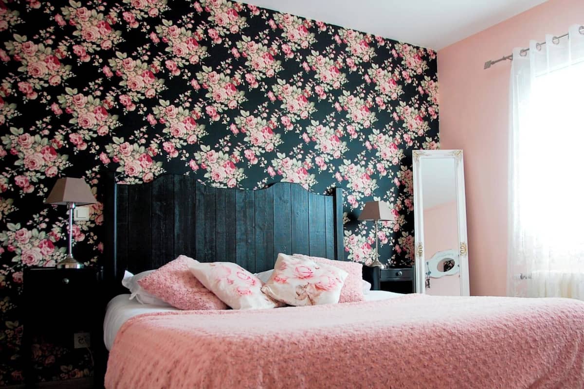 Quarto com papel de parede floral, cama com roupa de cama rosa, duas luminárias de cabeceira e um espelho na parede.