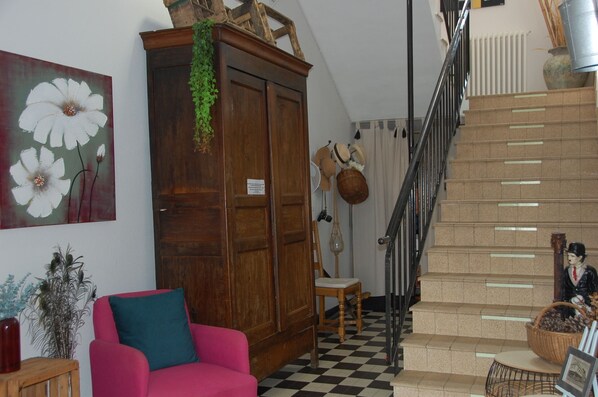 Un escalier avec un sol à carreaux, une armoire en bois ornée d’un élément décoratif vert suspendu, une chaise rouge et un mur sur lequel est accrochée une grande peinture de fleurs.