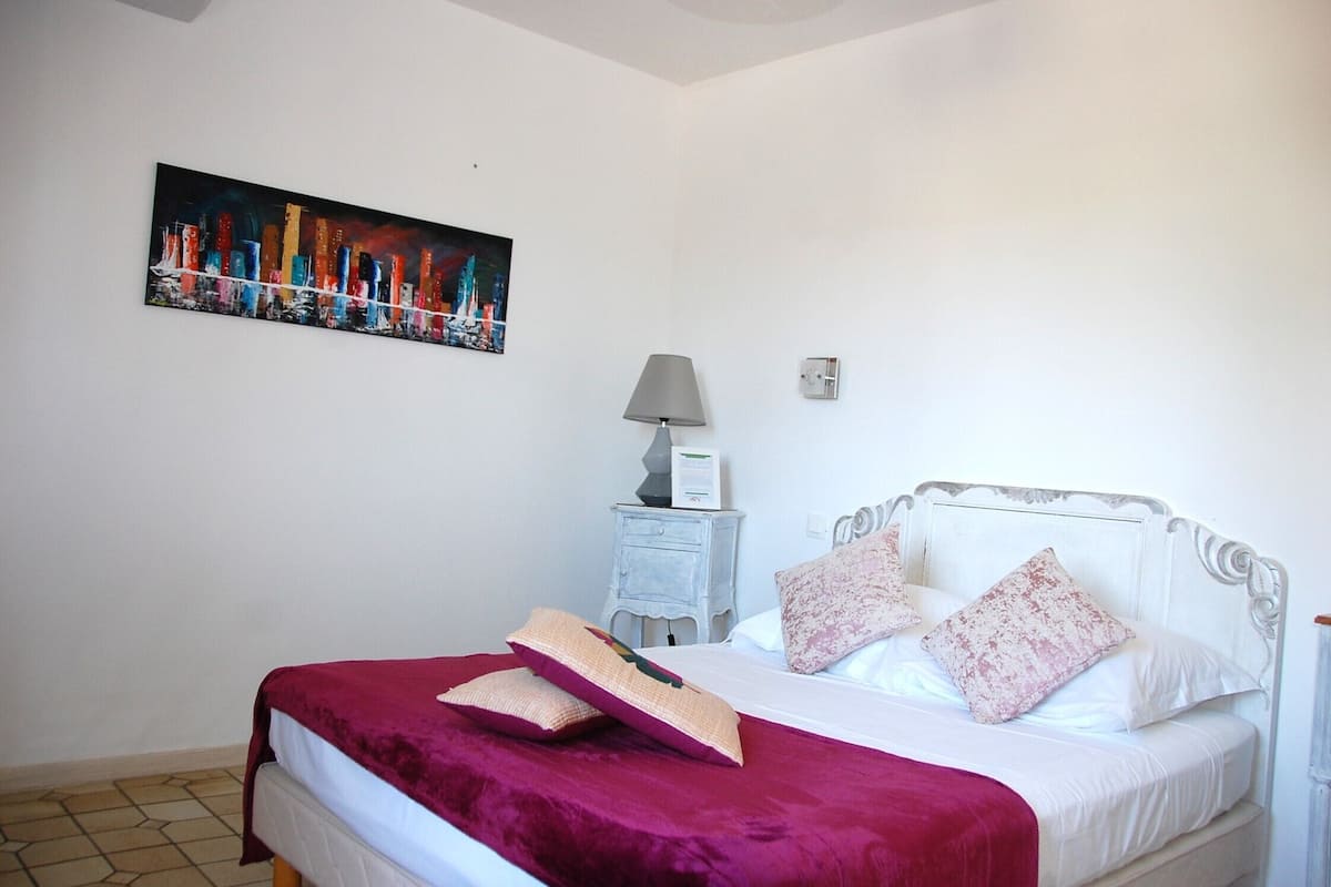 Quarto com cama, mesa de cabeceira, abajur e um quadro na parede.