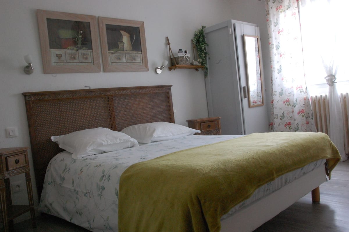 Quarto com cabeceira de madeira, cama com roupa de cama branca e manta verde, mesa de cabeceira, cômoda e espelho.