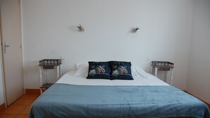 A neatly made bed with a blue blanket and floral pillows, a white wall with two wall-mounted nightstands, and a door on the left. - Le Pont de la Loire (Bourbon-Lancy)