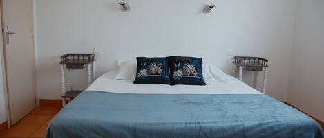 A neatly made bed with a blue blanket and floral pillows, a white wall with two wall-mounted nightstands, and a door on the left.