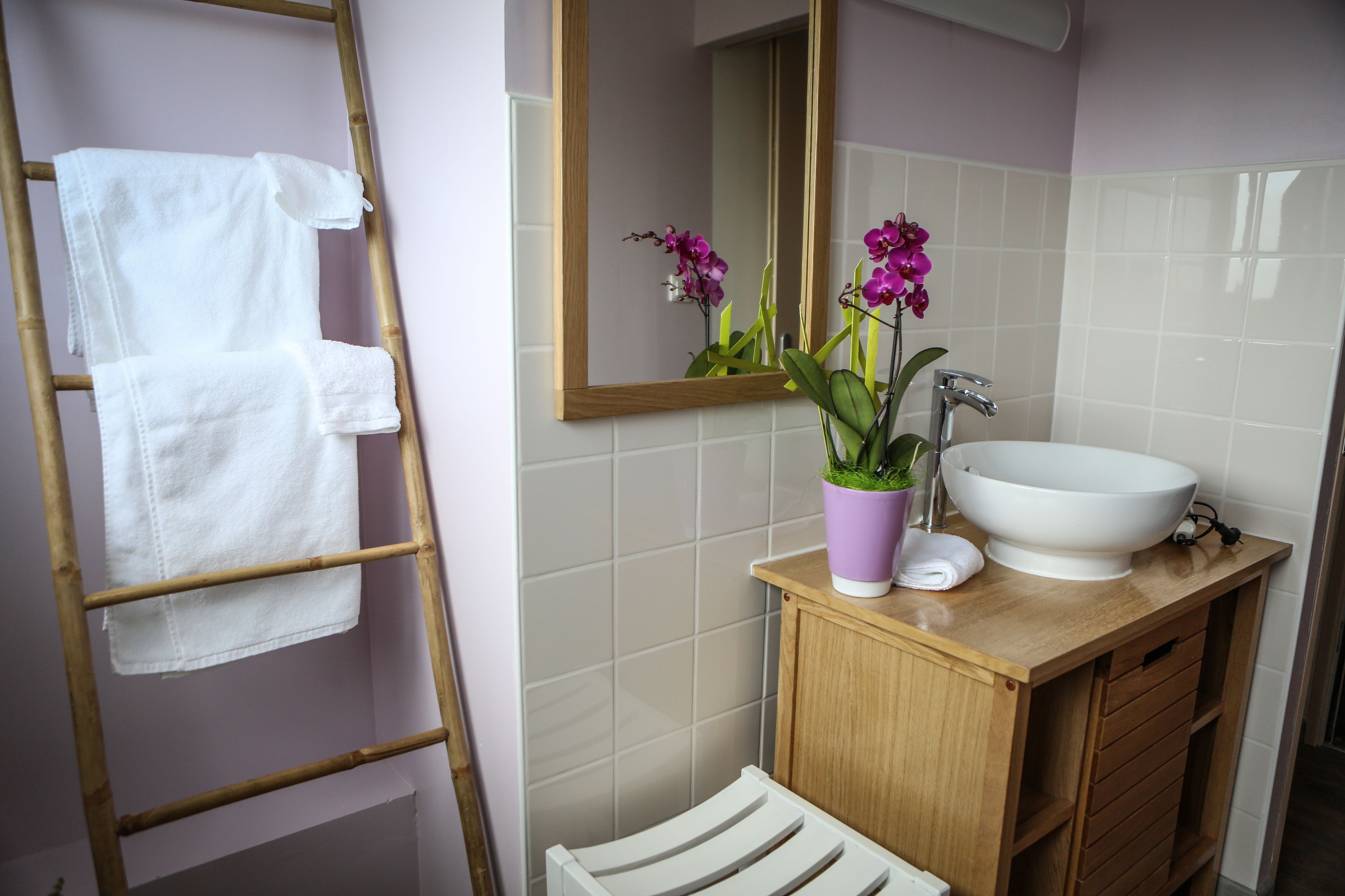 A bathroom with a wooden vanity, white sink, and a mirror. There is a towel rack with white towels and a potted orchid plant on the vanity.