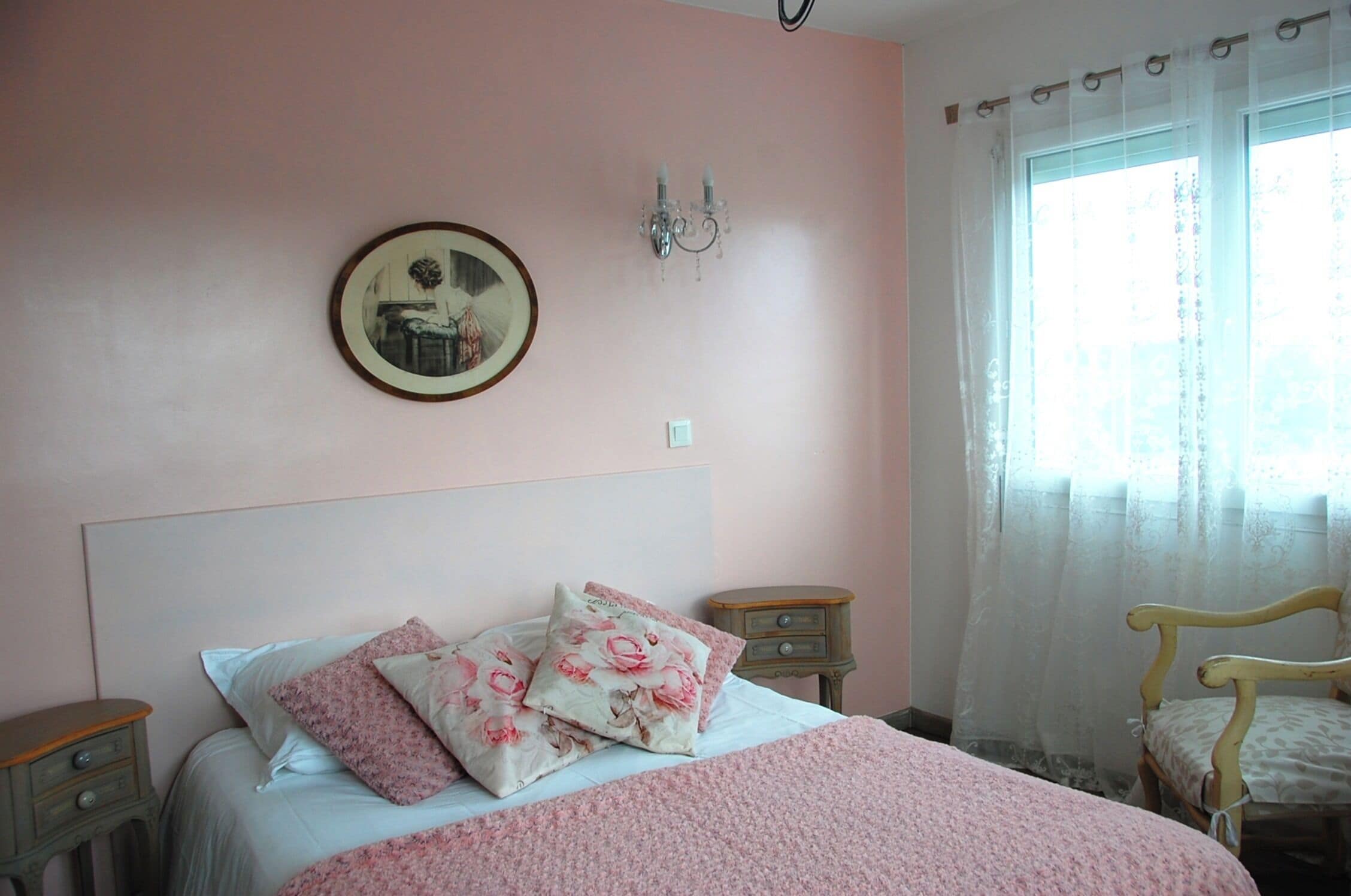 Un dormitorio con cama, almohadas, una mesita de noche, una silla, una lámpara de pared y un ventanal con cortinas.