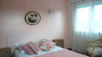 Un dormitorio con cama, almohadas, una mesita de noche, una silla, una lámpara de pared y un ventanal con cortinas.