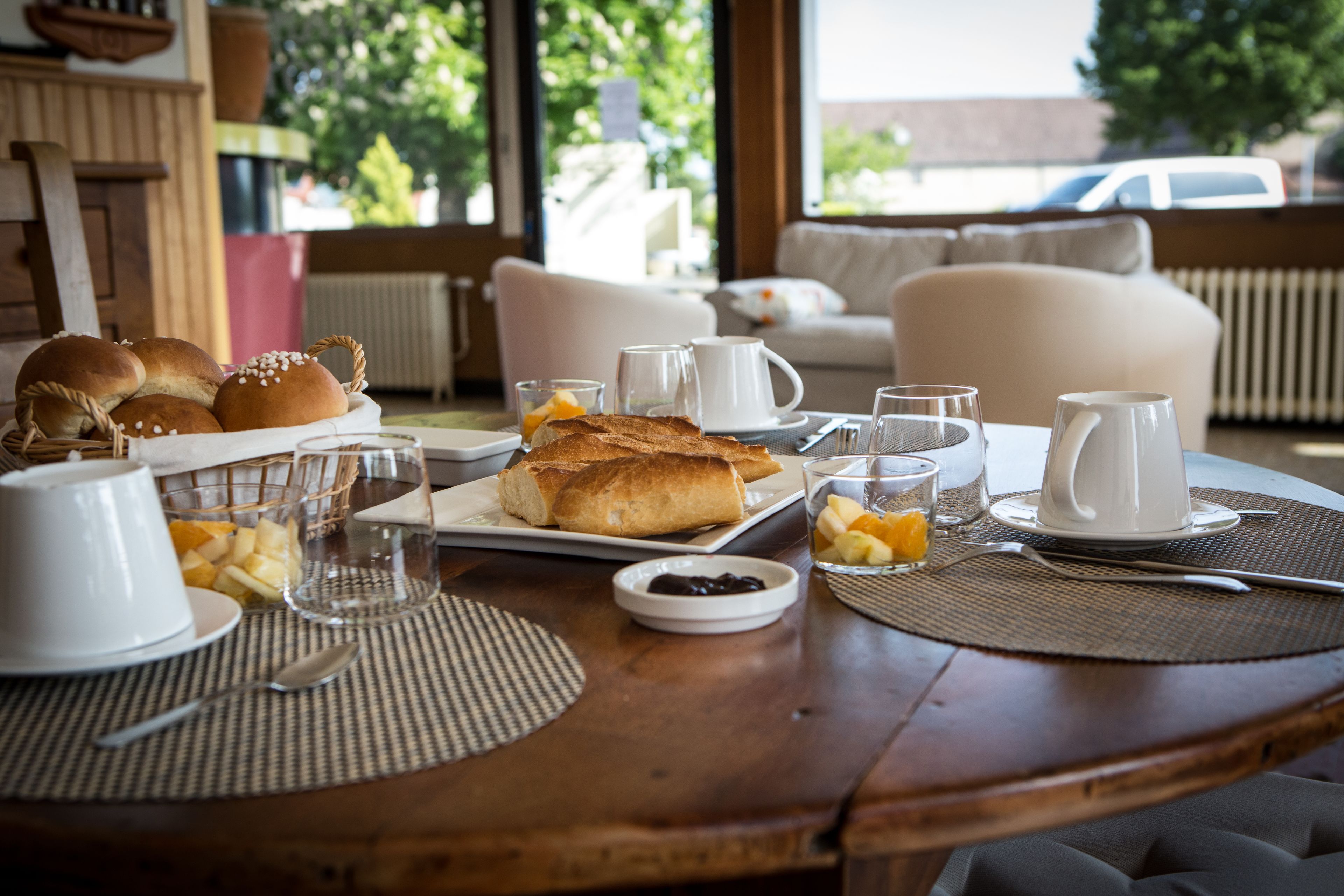 A breakfast table set with bread, fruits, and beverages, with a view of a car and greenery outside.