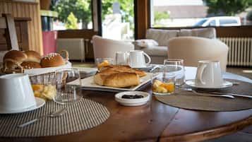 A breakfast table set with bread, fruits, and beverages, with a view of a car and greenery outside.