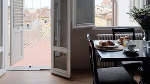 Un balcón abierto con una mesa dispuesta para el desayuno, incluyendo croissants y una taza de café, con vistas a un paisaje urbano con edificios históricos.