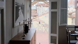 Una habitación con un armario de madera, una mesa con sillas, un ventanal con vistas a edificios y una puerta que da al exterior.