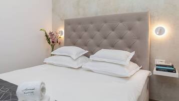 A neatly made bed with white linens and pillows, a tufted headboard, a bedside table with books, and a vase with flowers.