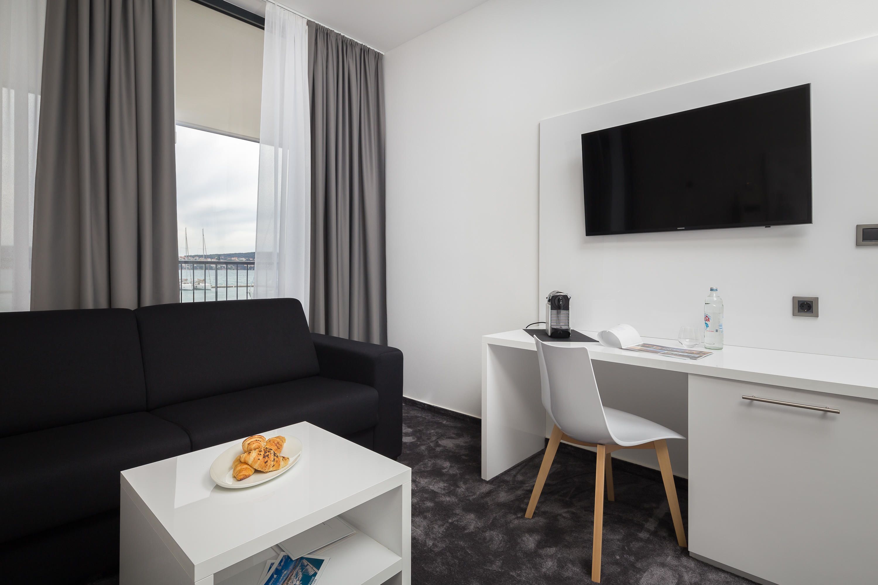 A modern hotel room with a black sofa, a white desk, a flat-screen TV, and a view of the sea through large windows.