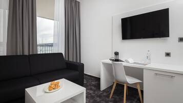 Quarto de hotel moderno com sofá preto, escrivaninha branca, TV de tela plana e vista para o mar através de grandes janelas.
