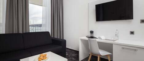 A modern hotel room with a black sofa, a white desk, a flat-screen TV, and a view of the sea through large windows.