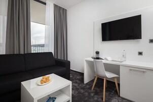 Quarto de hotel moderno com sofá preto, escrivaninha branca, TV de tela plana e vista para o mar através de grandes janelas.
