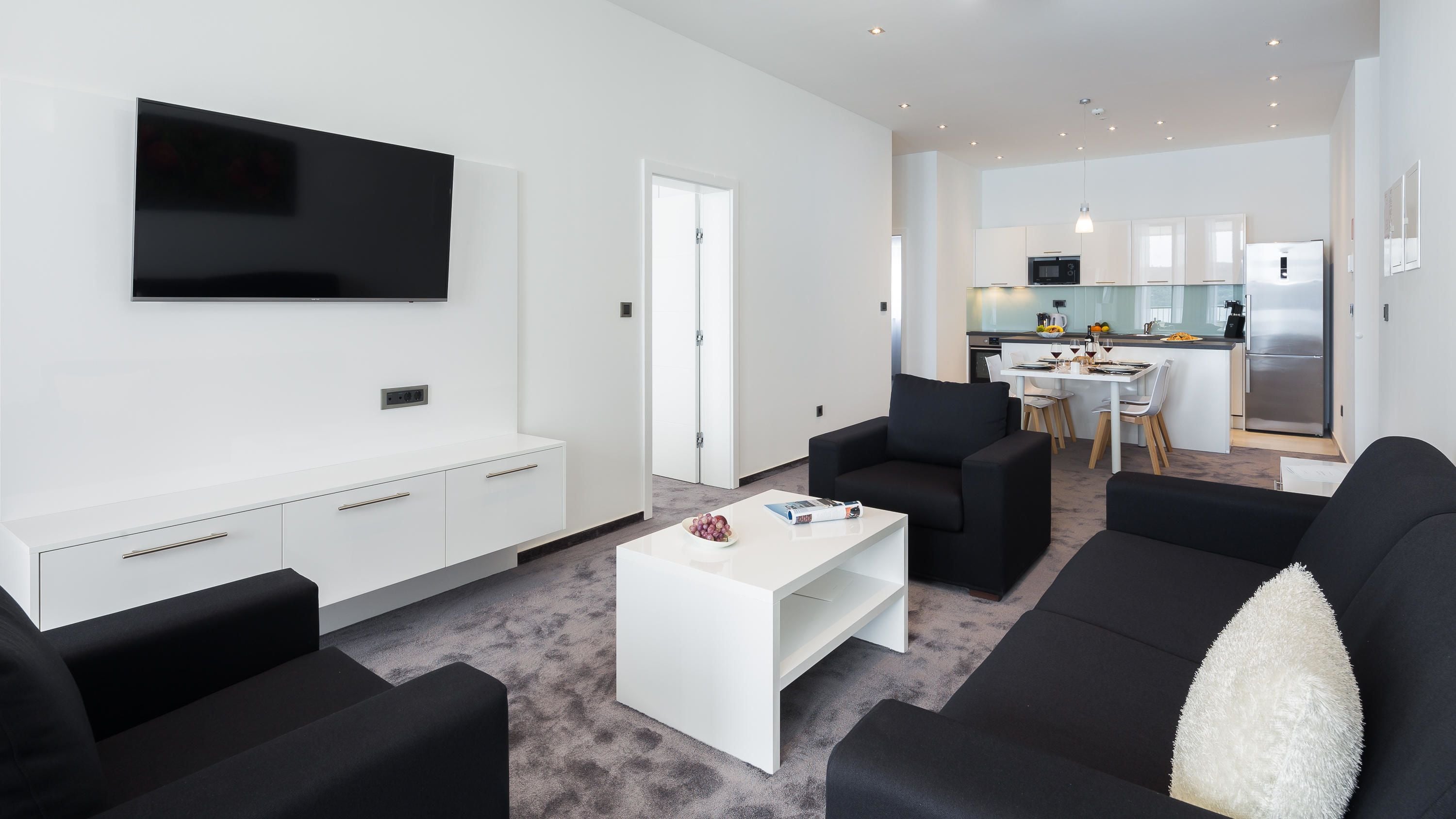 A modern living room with a flat-screen TV, black sofas, a white coffee table, and a dining area with a kitchen in the background.