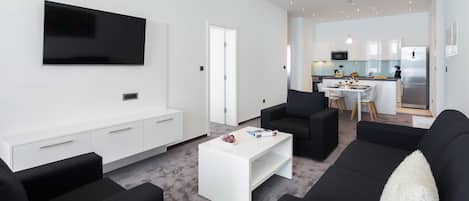 A modern living room with a flat-screen TV, black sofas, a white coffee table, and a dining area with a kitchen in the background.