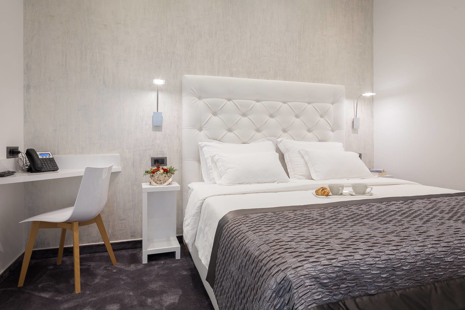 Ein modernes Hotelzimmer mit einer gepolsterten Kopfteil, einem weißen Schreibtisch mit Telefon, einem Sessel und einem Nachttisch mit Blumenarrangement.