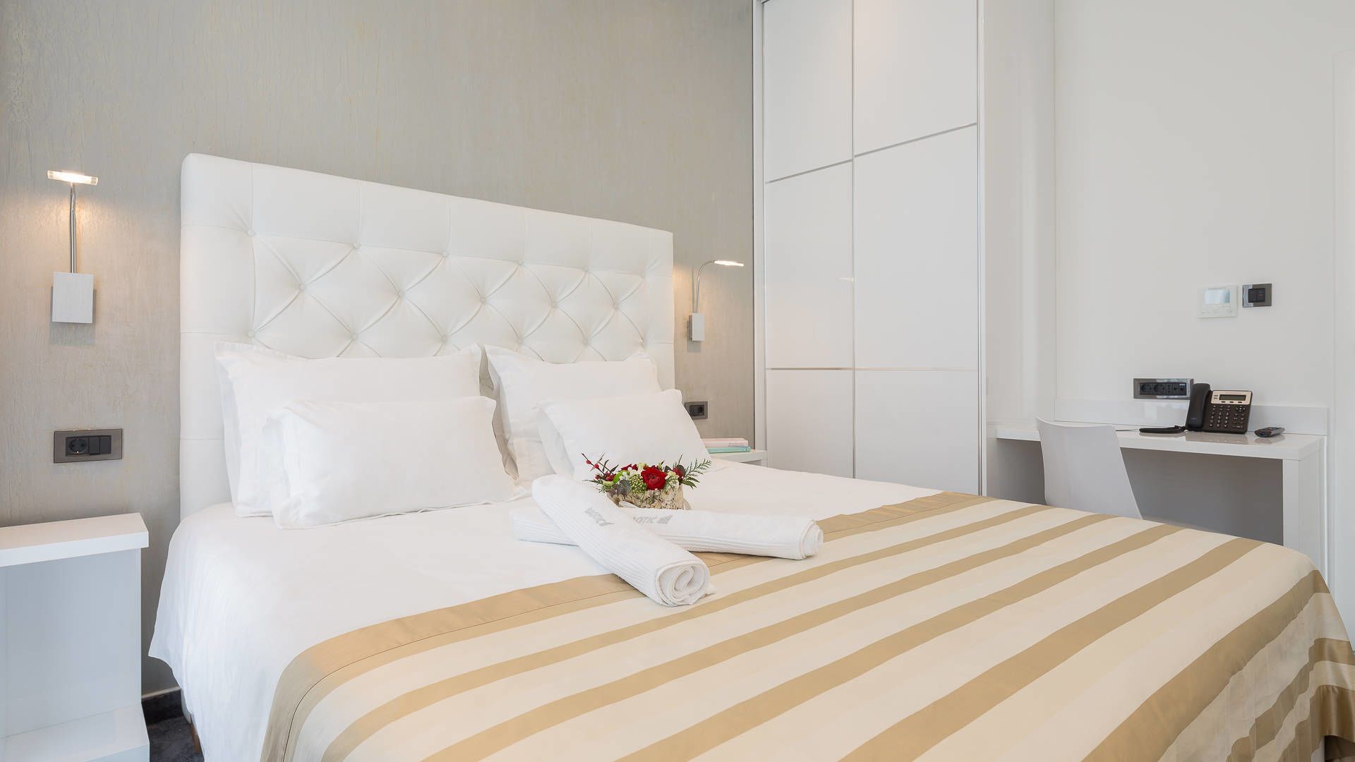 A hotel room with a large bed, white bedding, a striped bedspread, a bedside table with a lamp, and a phone on a desk.