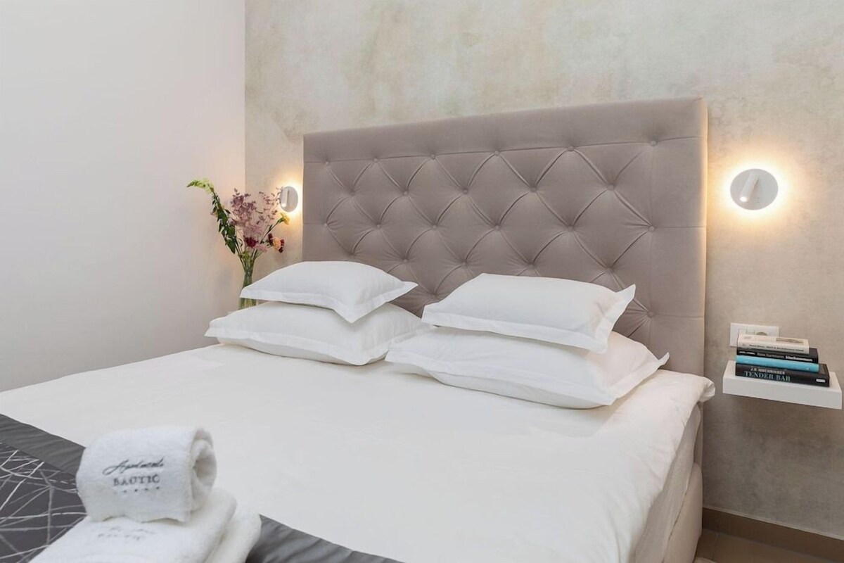 Un lit bien fait, avec une tête de lit capitonnée, une table de chevet avec une lampe et une petite étagère remplie de livres.