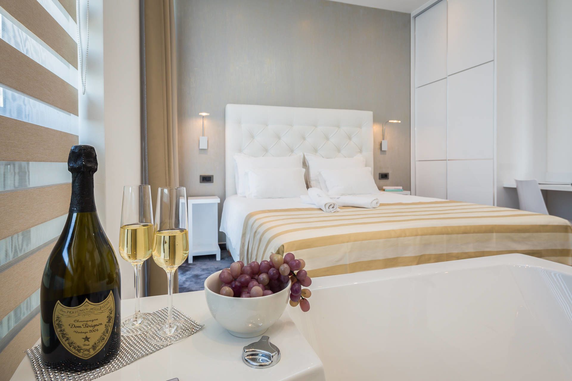 Habitación de hotel con cama, mesita de noche con una botella de champán, dos copas y un cuenco de uvas.