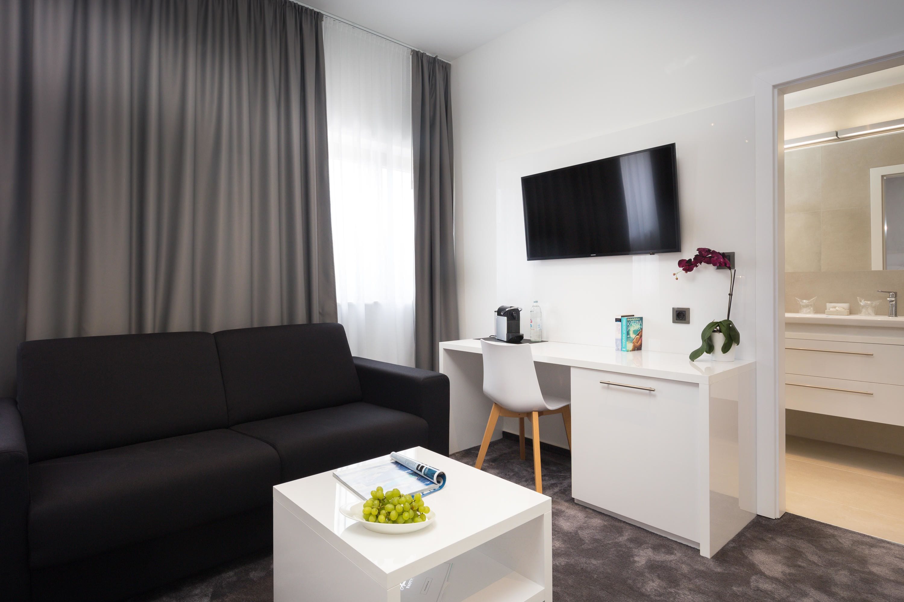 Una habitación de hotel moderna con un sofá negro, un escritorio blanco, un televisor de pantalla plana y una pequeña mesa con un plato de uvas.