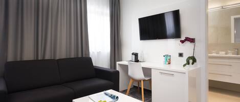 Une chambre d’hôtel moderne avec un canapé noir, un bureau blanc, un téléviseur à écran plat et une petite table sur laquelle se trouve une assiette de raisins.
