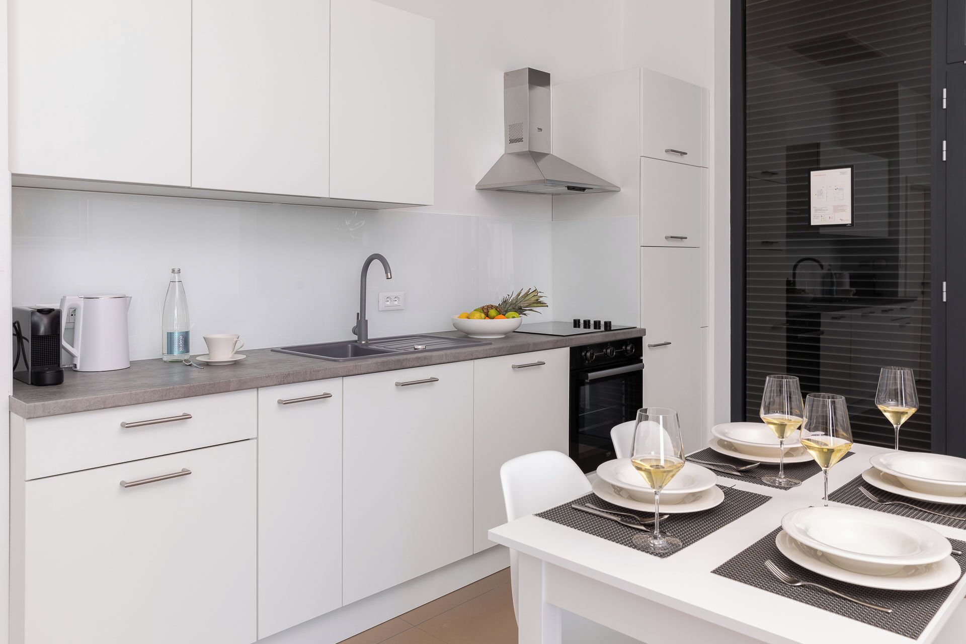 Cuina moderna amb armaris blancs, campana extractora d'acer inoxidable i zona de menjador preparada per a dinar, amb plats i gots blancs.